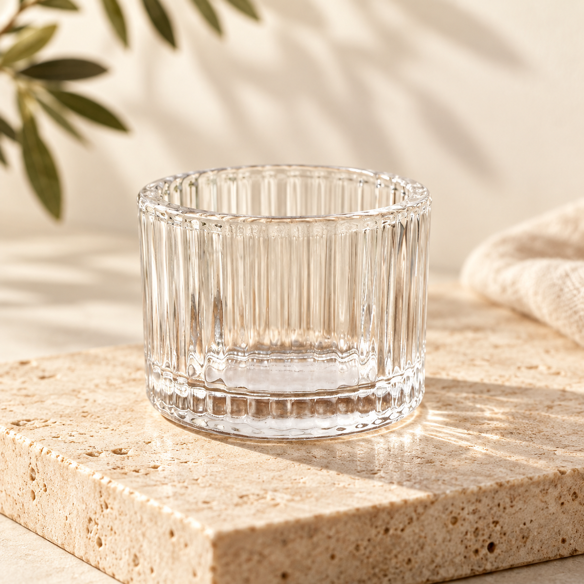 Empty clear ribbed glass tea light holder styled on warm stone with ivory linen and olive greenery.